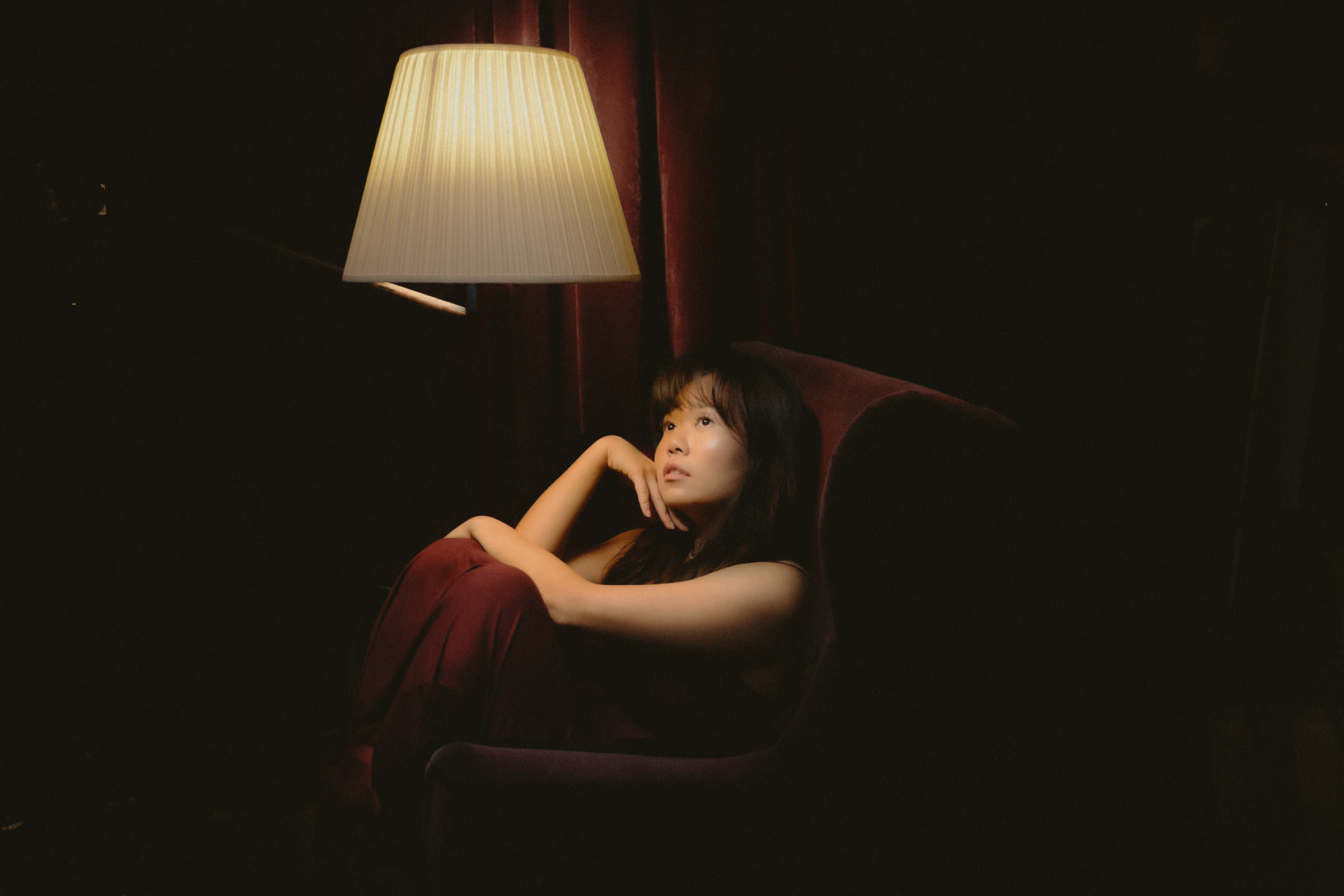 elysrei in a red velvet armchair under a lamp, looking up